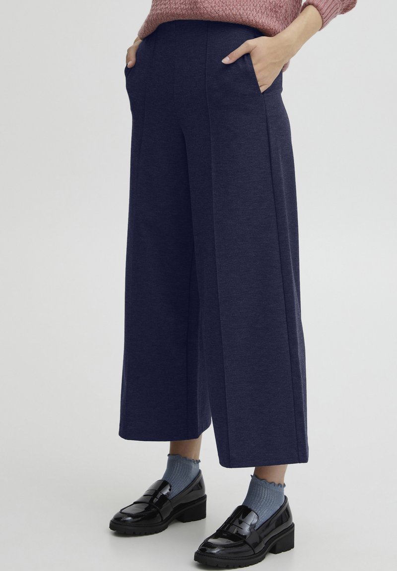 Marineblauwe wijde broeken met twee zijzakken, gemaakt van een zachte, gestructureerde stof, gecombineerd met zwarte instapschoenen en blauwe geribbelde sokken.