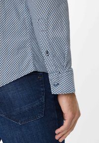Camicia a maniche lunghe con motivo a diamante blu e bianco, con polsini a un bottone e tessuto testurizzato. Indossata con jeans scuri.