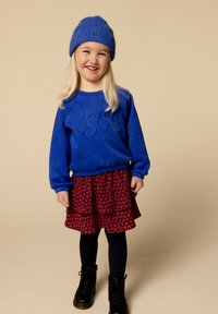 Blauwe sweatshirt met gestructureerde afwerking en de tekst "Amma", gecombineerd met een rode bloemenrok in tiers, zwarte panty’s en zwarte laarzen.