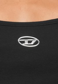 Čierna rebrovaná tkanina so širokým výstrihom; uprostred má strieborný oválny logo akcent. Textúra pôsobí mäkko a priliehavo.