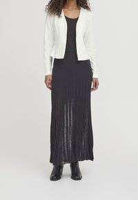 Bílá padnoucí bunda s dlouhými rukávy přes černý tílko a dlouhou černou plisovanou sukni, doplněná černými kotníkovými botami.