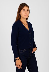Cardigan corto blu navy in maglia strutturata, con scollatura a V, maniche lunghe e cinque bottoni frontali. Jeans in denim scuro completano il look.