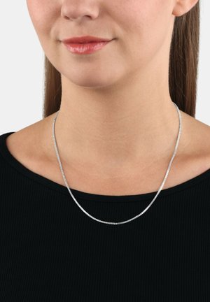 Primo piano di una donna che indossa una delicata collana di argento su una maglietta nera a costine, con i capelli castani lisci in vista.