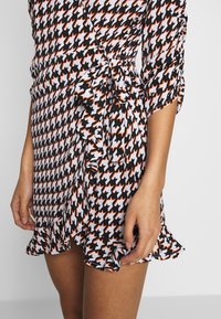 Läheltä kuvattu nainen, joka on pukeutunut musta, valkoiseen ja oranssiin houndstooth-kuvioiseen kietaisupukuun, jonka helma on frillalla ja vyötäröllä on solmittu nauha.