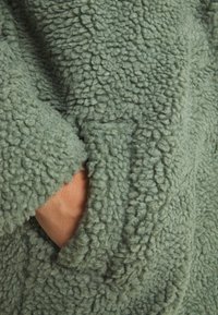 Grön fleecejacka med en texturerad, plysch yta; har en sidficka som delvis är synlig och visar det mjuka, varma materialet.