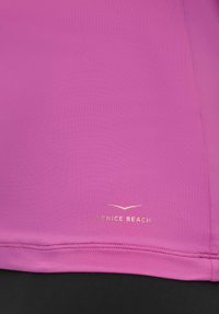 Pinkfarbener Sporttop mit glattem Stoff, figurbetontem Design und einem goldfarbenen "VENICE BEACH"-Logo am Saum, das einen Kontrast zum darunterliegenden schwarzen Material bildet.
