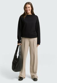 Sweatshirt noir à manches longues avec un col bateau, associé à un pantalon beige à jambes larges. Le mannequin tient un grand sac en cuir noir à bandoulière.