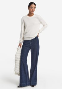 Weißer Pullover, weit geschnittene dunkle Denim-Jeans und schwarze spitze Schuhe. Das Modell hält eine cremefarbene Daunenjacke. Einfaches, klassisches Design und Farben.