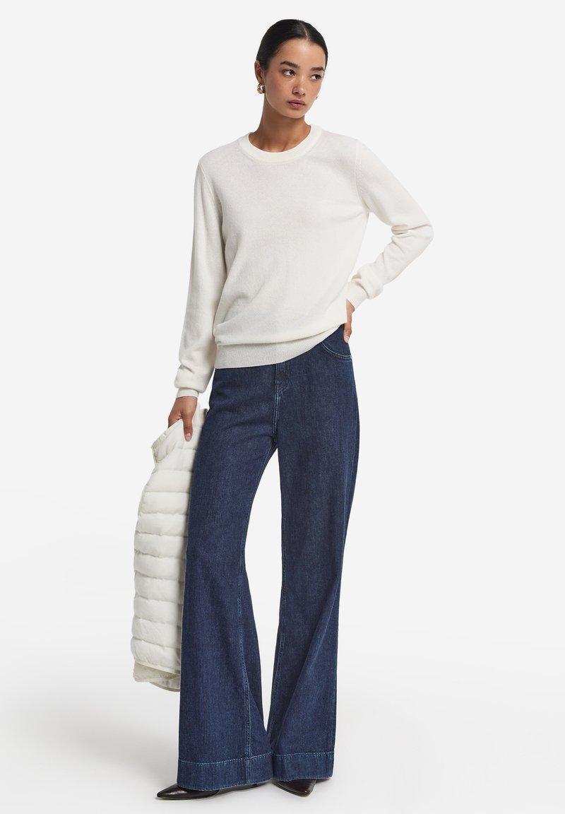 Weißer Pullover, weit geschnittene dunkle Denim-Jeans und schwarze spitze Schuhe. Das Modell hält eine cremefarbene Daunenjacke. Einfaches, klassisches Design und Farben.