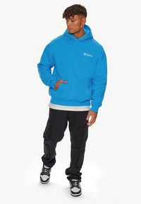 Blaues Kapuzen sweatshirt mit einer Fronttasche, das ein weißes Logo auf der Brust trägt. Getragen mit schwarzen Cargo-Hosen und mehrfarbigen Turnschuhen.