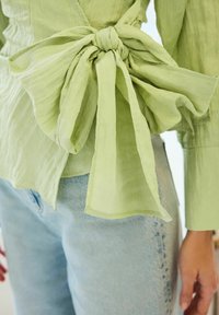 Ljust grön texturerad blus med en stor rosett på baksidan, i kombination med ljusblå denim. Tyget ser mjukt och lätt skrynkligt ut.
