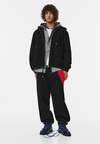 Giacca nera trapuntata con cappuccio, indossata sopra una camicia a quadri grigia e una t-shirt bianca, abbinata a pantaloni neri larghi con un accento rosso. Stivali neri.