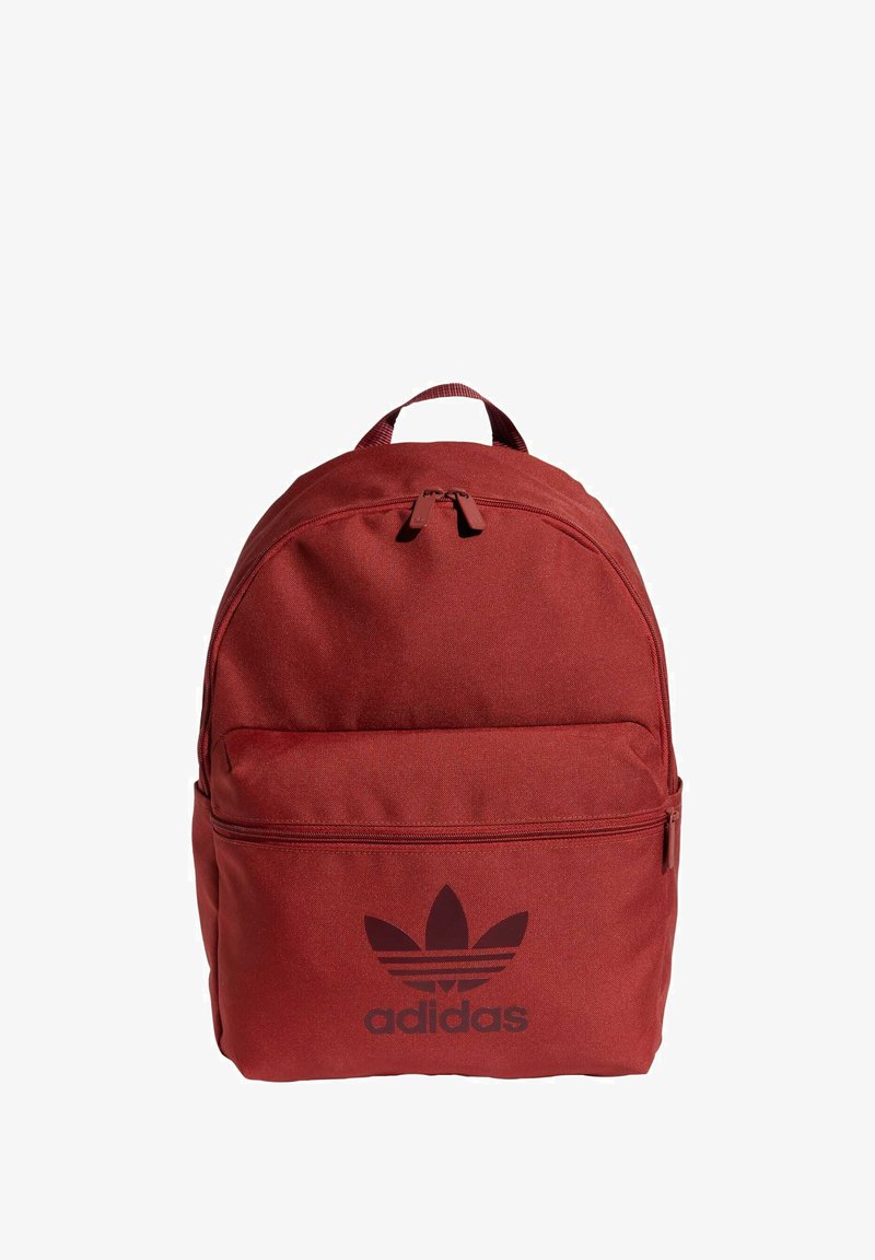 Punainen kangaskassi, jossa on pyöreä yläosa, etutasku vetoketjulla ja musta Adidas-logo edessä. Säädettävät olkaimet.