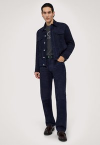 Marinblå mockajacka och byxset med två framfickor, tryckknappar och en strukturerad yta, kombinerat med en grå skjorta och bruna skor.