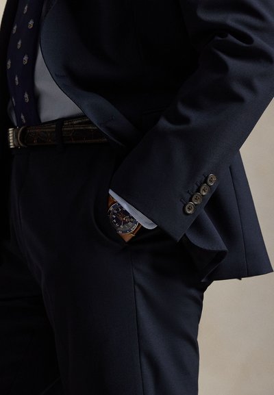 Ναυτικό κοστούμι με υφή, πουκάμισο με ρίγες και γραβατα με μοτίβο. Ρολόι με μπλε καντράν και καφέ δερμάτινο λουράκι διακρίνεται από την τσέπη.