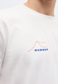 Chemise blanche avec le texte bleu "Mammut" et un graphique de montagne à contour orange sur la poitrine, portée par une personne debout.