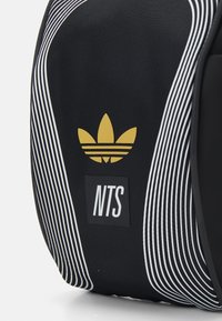 Bolsa negra con rayas blancas y un logo dorado de trebol de Adidas. Presenta un parche rectangular "NTS", elaborado con material duradero.