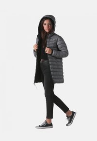 Manteau gris rembourré avec capuche, doté d'un design matelassé, de manches longues et d'une fermeture éclair. Porté sur un jean noir et des baskets.