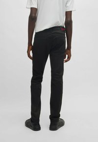 Jeans noirs à coupe slim, dotés d'un design classique à cinq poches et d'une étiquette de logo rouge sur la taille, portés avec des chaussures noires.