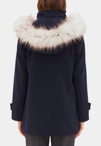 Woman wearing a navy blue coat with a large beige faux fur-trimmed hood, shown from the back against a plain background.