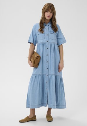 Nő világoskék gombos farmer midi ruhában, barna borítéktáskával a kezében, barna mokaszin cipőben, egyszínű háttér előtt.