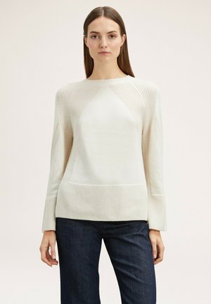 Crèmekleurige gebreide trui met een ribbelstructuur, ronde halslijn en licht uitlopende mouwen, gecombineerd met donkere jeans. Kenmerkt zich door een naadloos schouderontwerp.