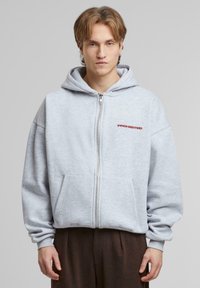 Hoodie zippé gris à coupe ample, avec deux poches avant et le texte "PROHIBÉ" en rouge. Texture douce, avec une grande capuche et des poignets côtelés.