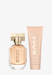 Botella de perfume de cristal en forma geométrica con tapa dorada, junto a un tubo de loción rosa con texto en relieve. Ambos productos etiquetados como "BOSS."