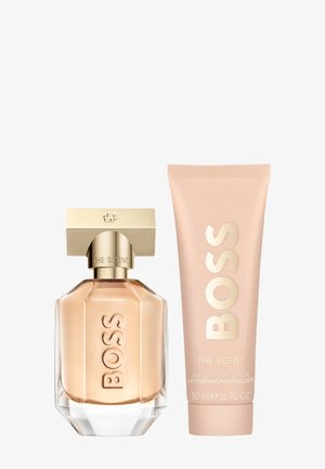 Glass perfume bottle in a geometric shape with a gold cap, next to a pink lotion tube featuring embossed text. Both products labeled "BOSS."