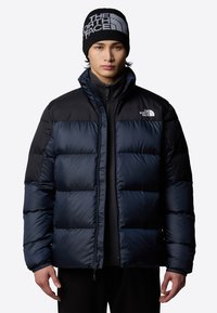 Marineblå puffjacka med quiltat mönster och svarta ärmar. Bärs över en svart zip-up. Har The North Face-logotypen på bröstet och en mössa.