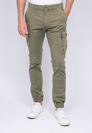 Homme portant un pantalon jogger cargo vert olive avec des poignets élastiques, des poches avant et une poche latérale zippée, assorti à des baskets blanches.