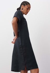 Leinenkleid in Dunkelblau, mit kurzen Ärmeln, einem seitlichen Schlitz und einer Brusttasche. Der Stoff hat eine glatte Textur und einen lockeren Schnitt.