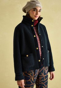 Námornícka fleecová bunda so zlatými gombíkmi a dvoma vreckami. Nosená nad červeným vzorovaným sveterom, kombinovaná so vzorovanými nohavicami a béžovou pletenou čiapkou.