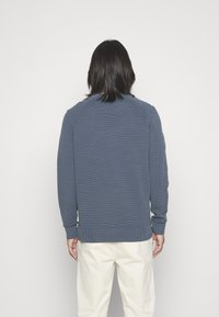 Blauer strukturierter Sweatshirt mit gerippten Bündchen und Saum, rundem Halsauschnitt und Raglanärmeln. Kombiniert mit beigen Hosen.