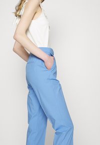 Vaaleansiniset housut, joissa on slim fit -leikkaus, sivutaskut ja siisti viimeistely. Yhdistetty valkoiseen yläosaan, jossa on röyhelövarret.