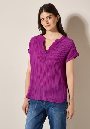 Haut à manches courtes, col en V, texturé en violet vif. Présente un design en tissu froissé et des fentes sur les côtés, associé à un jean bleu décontracté.