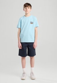 T-shirt di cotone azzurro chiaro con maniche corte e stampa grafica sul lato sinistro del petto, abbinata a pantaloni corti blu scuro e scarpe da ginnastica bianche.