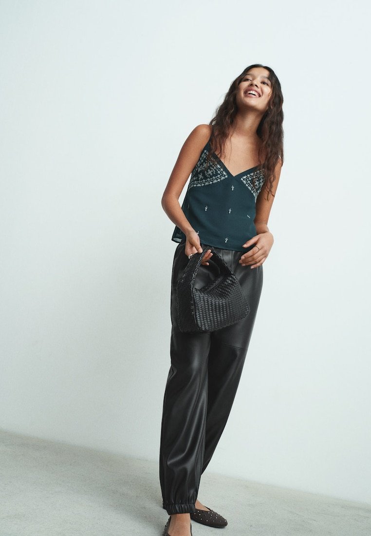 Tirkizna majica na bretele s čipkastim detaljima, kombinirana s crnim hlačama od umjetne kože i crnom pletenom torbom; model ima tamnu, valovitu kosu.