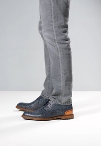 Scarpe in pelle blu testurizzata con motivo rettile, suola marrone e lacci decorativi, indossate con jeans grigi a taglio slim.