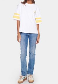 Osoba v bílém oversized tričku s žlutými pruhy na rukávech, modrých džínách a béžových teniskách se zelenými pruhy.