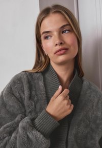 Šedý plyšový cardigan s ribbed límcem a manžetami, který má měkký, texturovaný povrch. Modelka drží límec blízko svého krku.