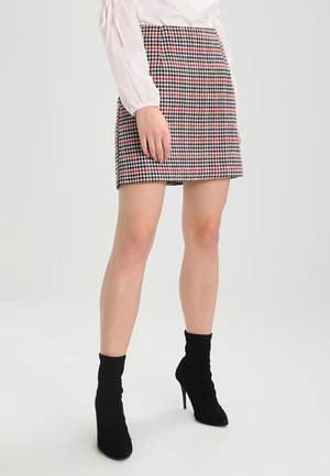 Kobieta ubrana w mini spódniczkę w czarno-biało-czerwoną kratę, białą bluzkę z długim rękawem oraz czarne skórzane botki na obcasie, stojąca.