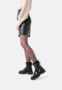 Botas de tobillo de cuero negro con un puño de peluche, detalles de hardware plateado y una suela gruesa. Llevadas con una falda de cuero negro y medias transparentes.