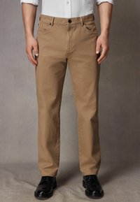 Pantalons en coton khaki avec une coupe droite, dotés de cinq poches, d'une fermeture à un bouton et d'une texture lisse ; portés avec des chaussures de ville noires.