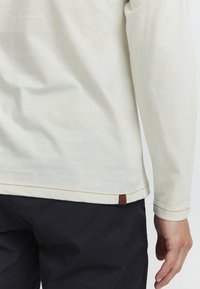 Camisa de manga larga color crema hecha de un tejido suave con un dobladillo redondeado, acentuada por costuras marrones y un pequeño parche de logo en la parte inferior.