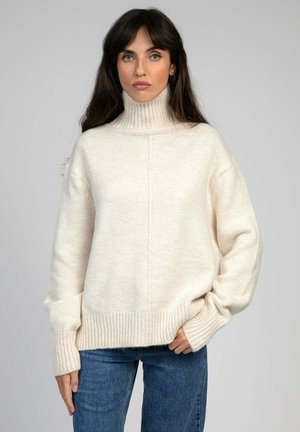 Femme aux longs cheveux foncés portant un pull à col roulé couleur crème et un jean bleu, debout devant un fond clair uni.