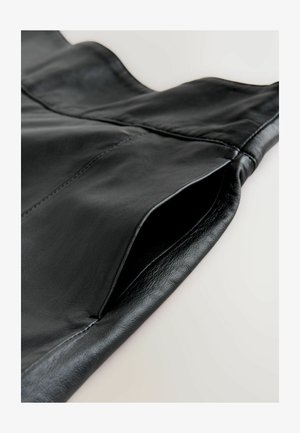 Schwarzes Lederkleidungsstück mit glatter Textur, strukturiertem Design und sichtbarem Taschenöffnung. Genähte Details entlang der Nähte.