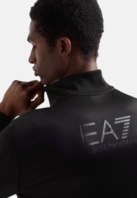 Sort, tætsiddende zip-jakke med høj krave og hævet EA7 Emporio Armani logo på ryggen. Glat tekstur og moderne design.