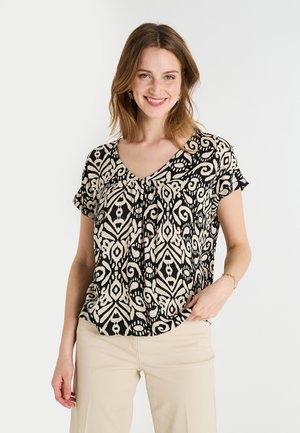 Blouse à manches courtes avec un motif tourbillonnant noir et crème, décolleté en V, et en tissu léger, associée à un pantalon beige.