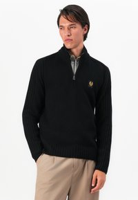 Homem com um suéter preto de gola alta com zíper, por cima de uma camisa cinza-clara, combinado com calças bege, de pé com uma mão no bolso.